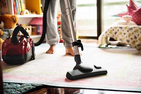 Person vacuuming a carpet in a brightly lit room with toys and a bed visible in the background.